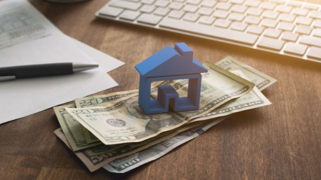 a-blue-home-figurine-resting-on-a-money-with-a-contract-in-the-background.png